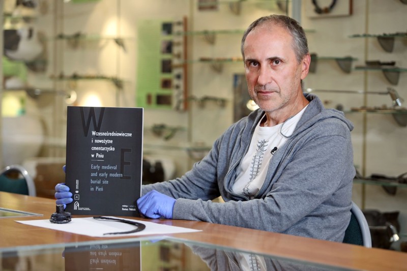 Professor Dariusz Poliński holds a publication on the Pień cemetery, with archaeological objects on the table.